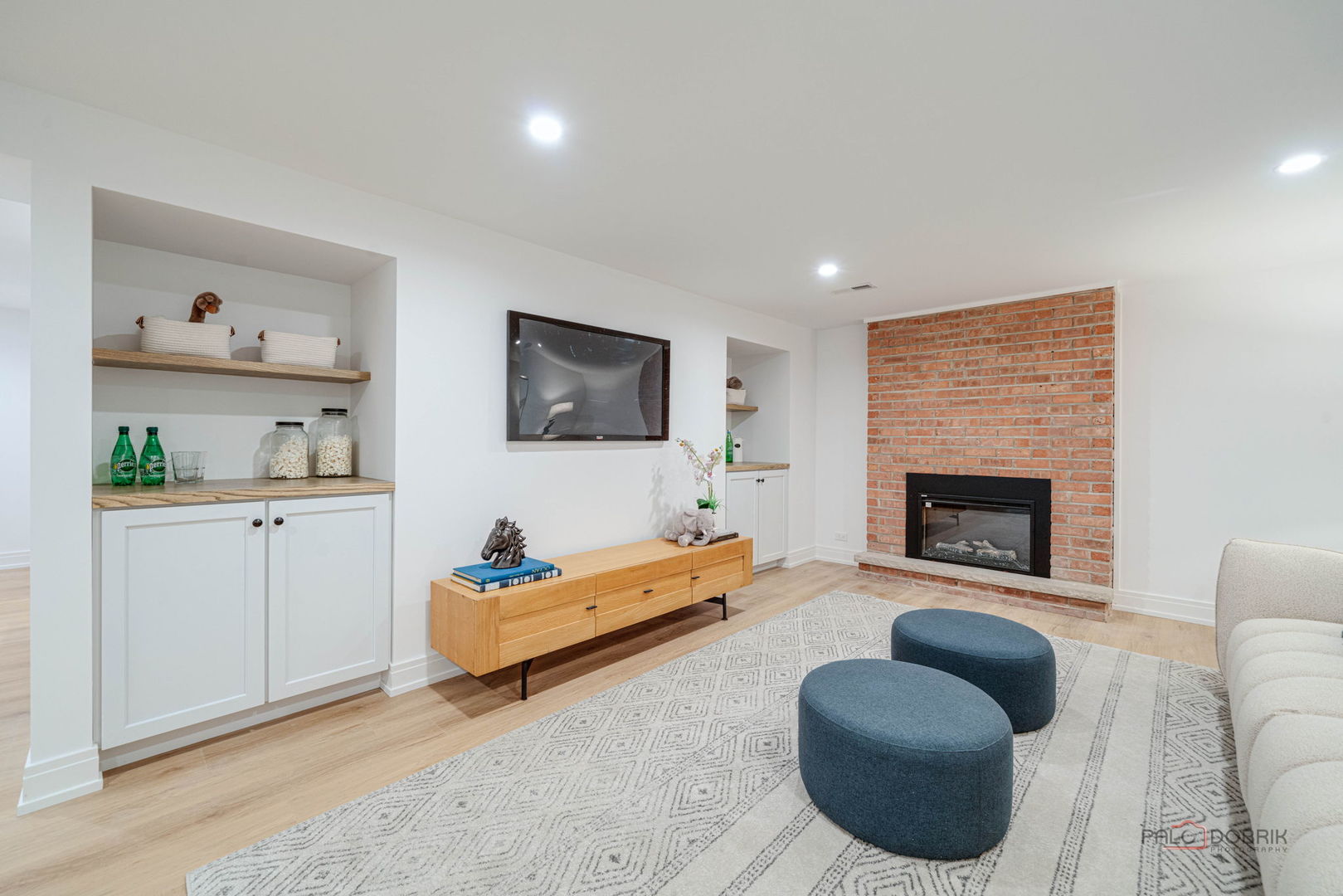 Living room with a brick fireplace, built-in shelving, and two blue ottomans on a patterned rug.