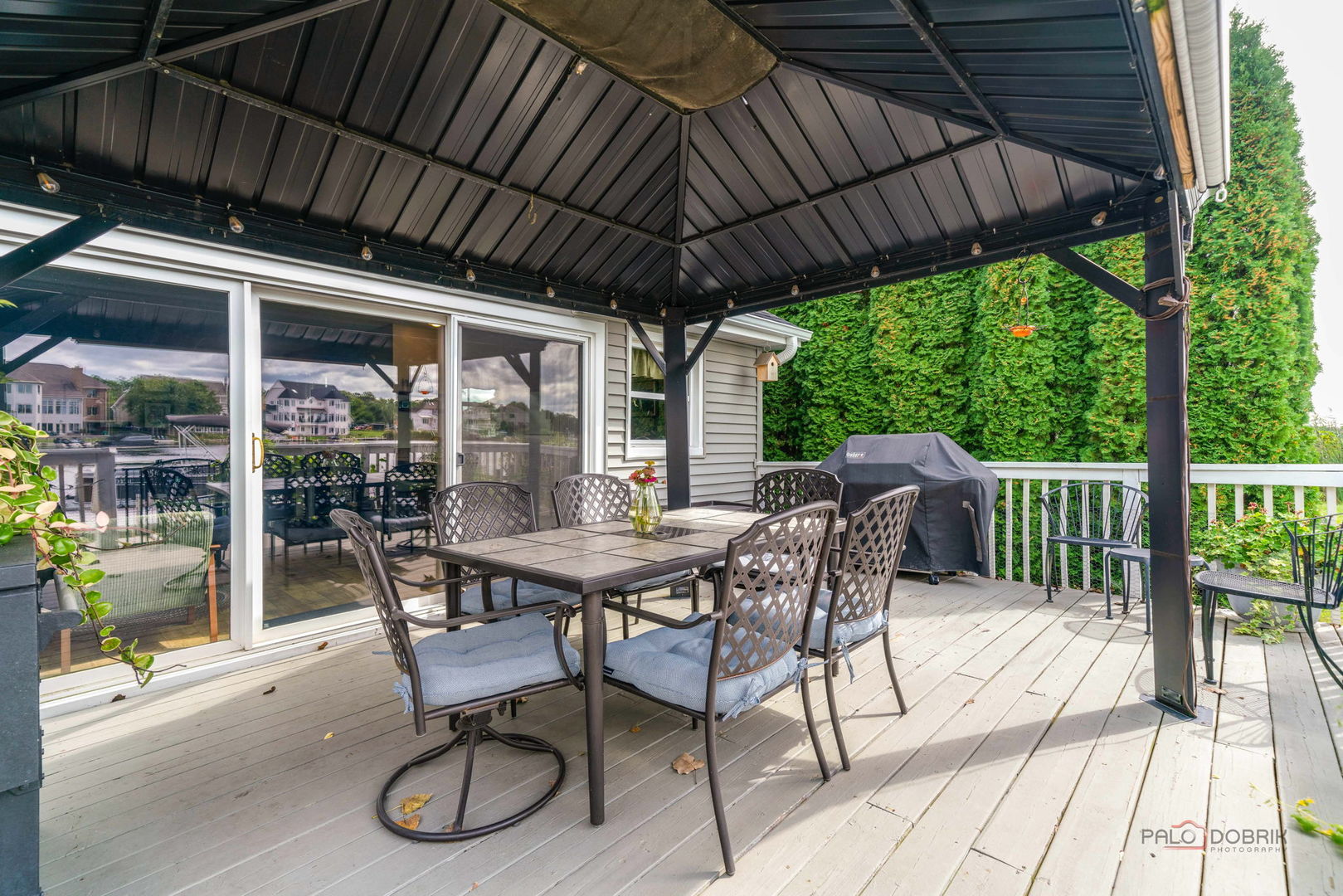 Covered deck with patio dining set, grill, and tall evergreen hedges.