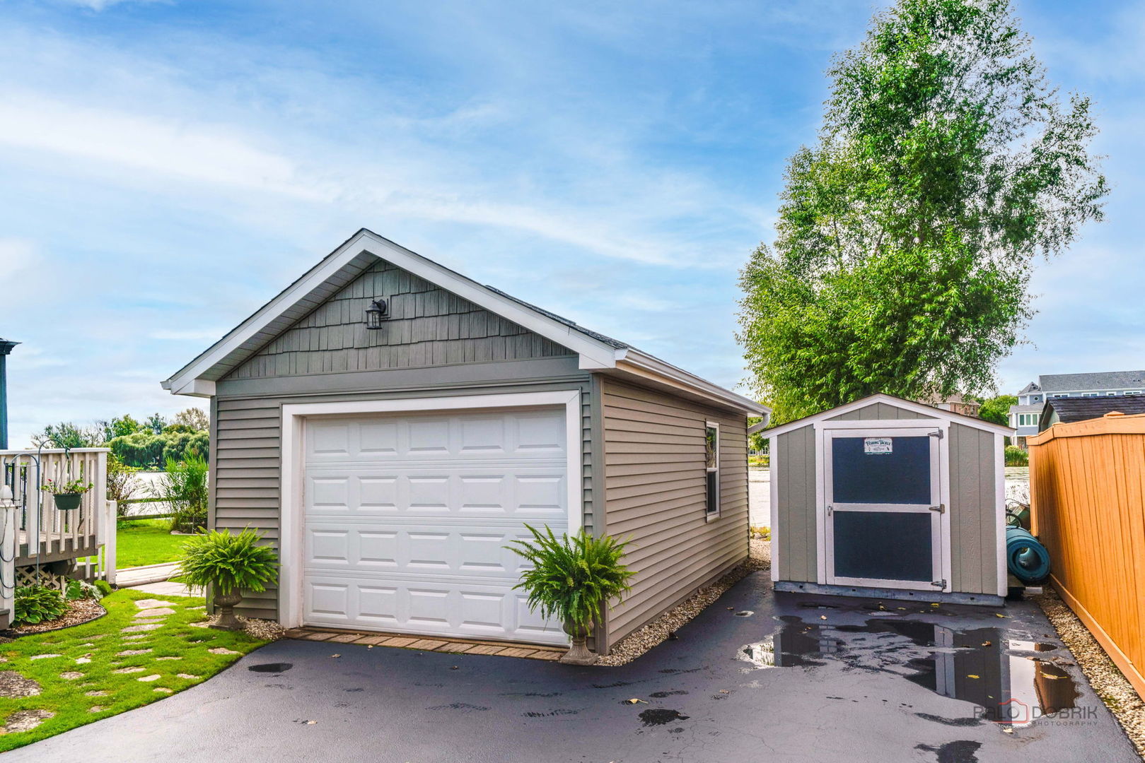 Detached gray garage with white overhead door, beside a matching storage shed and wet driveway.