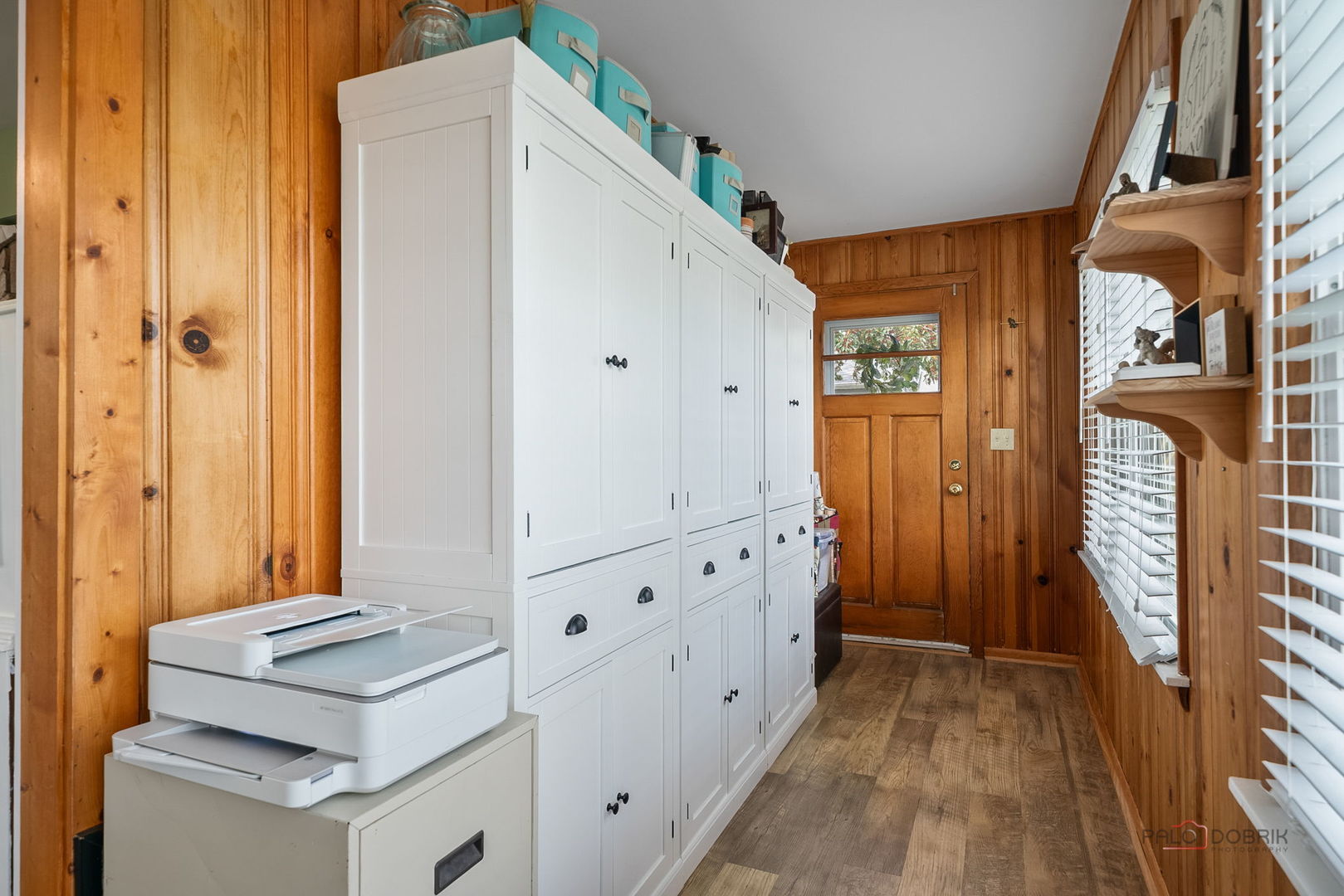 Wood-paneled storage room with white built-in cabinets, a printer, and a wooden exterior door.