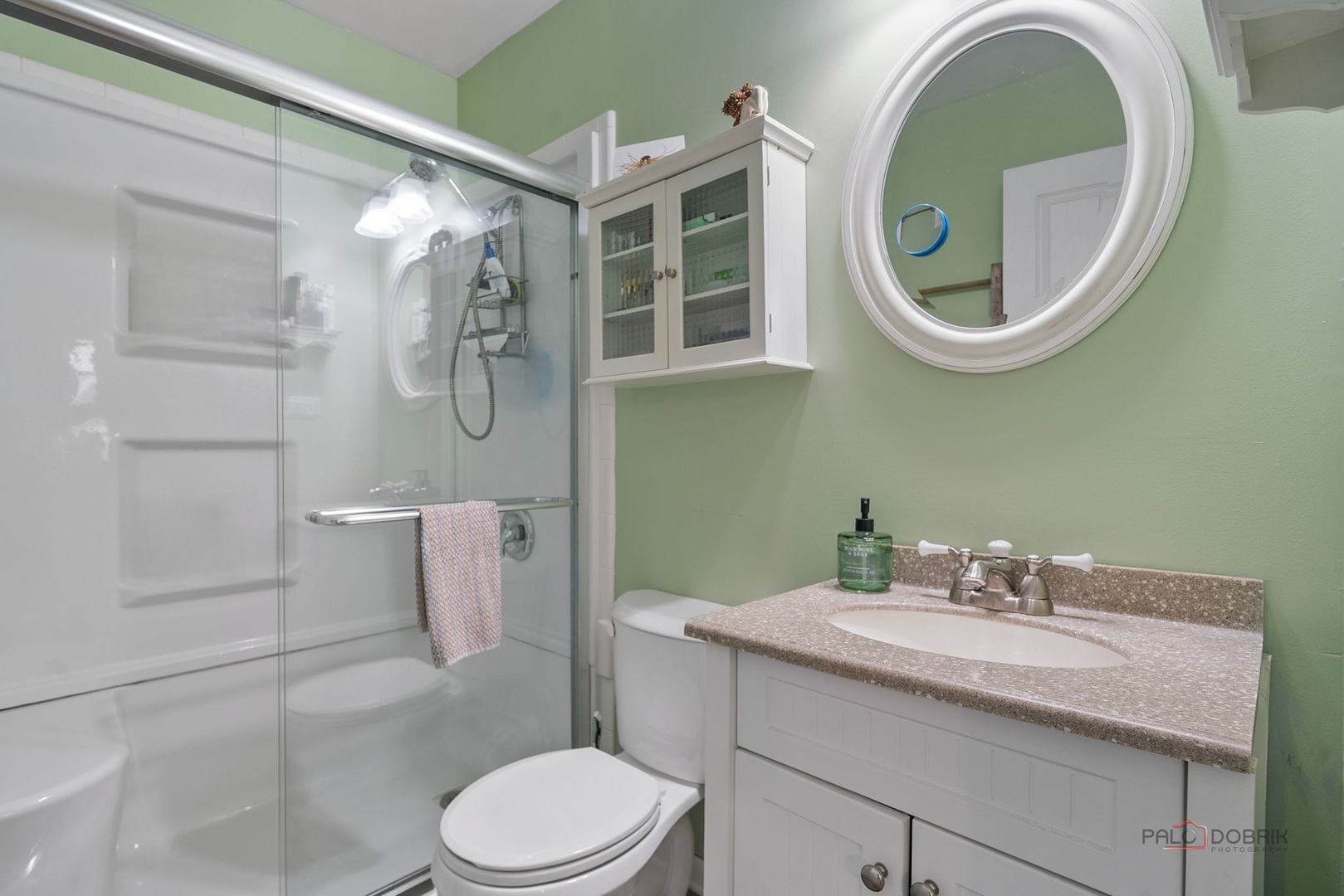 Bathroom with glass-enclosed shower, white vanity, round mirror, and pale green walls.