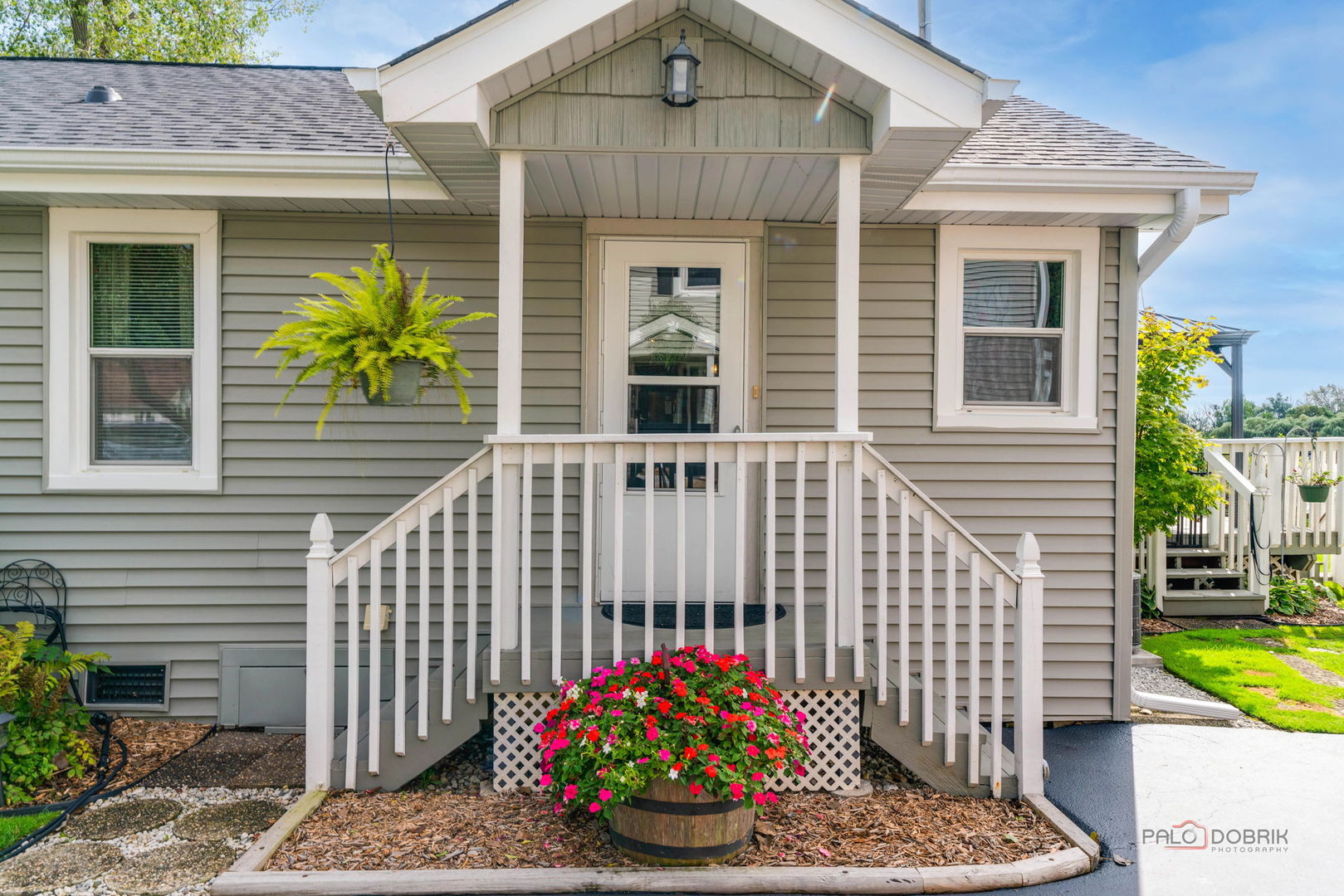Gray-sided home with a covered white entry, front steps, hanging fern, and bright flowers.