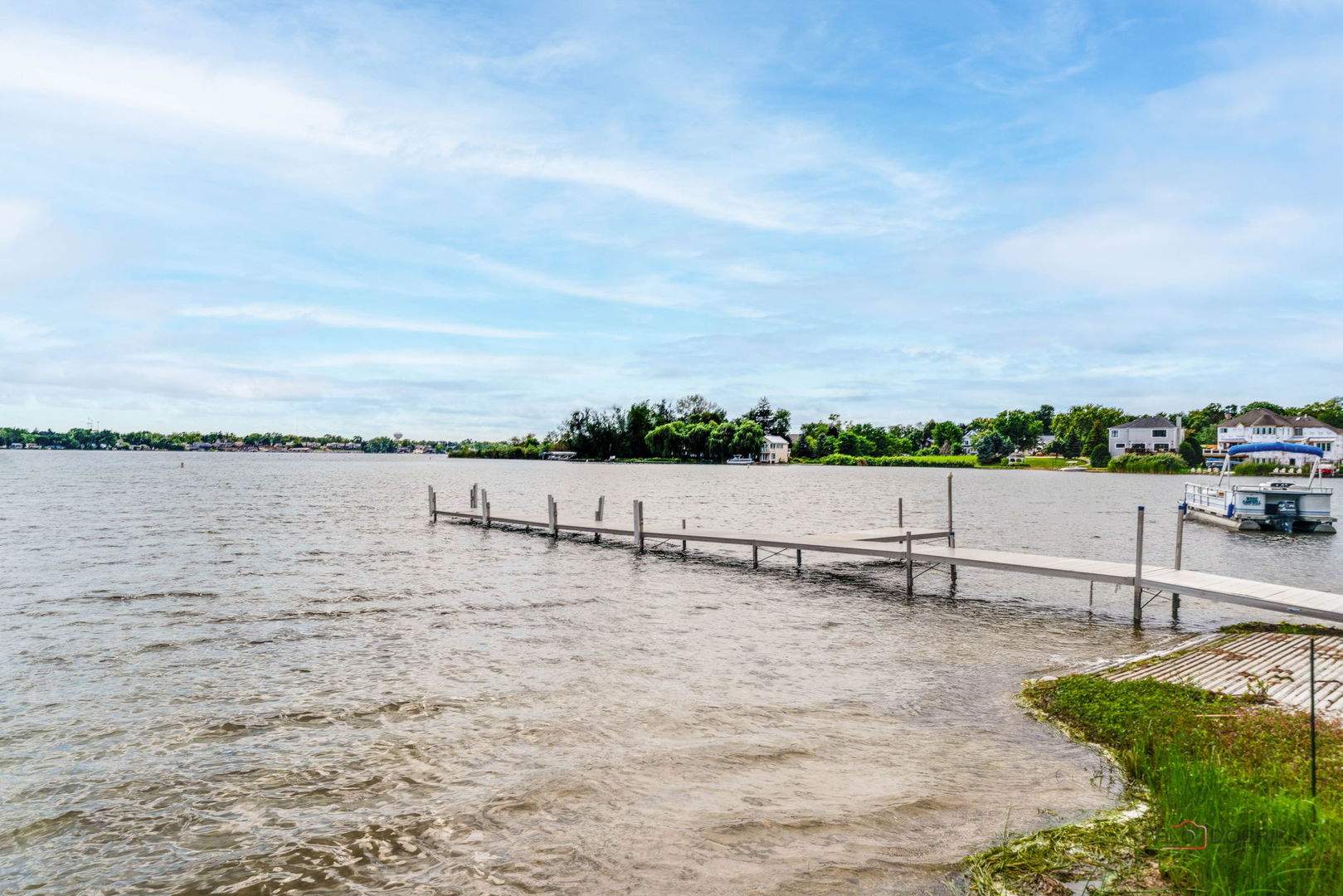 Lakefront view with a long dock, pontoon boat, and homes across the water.