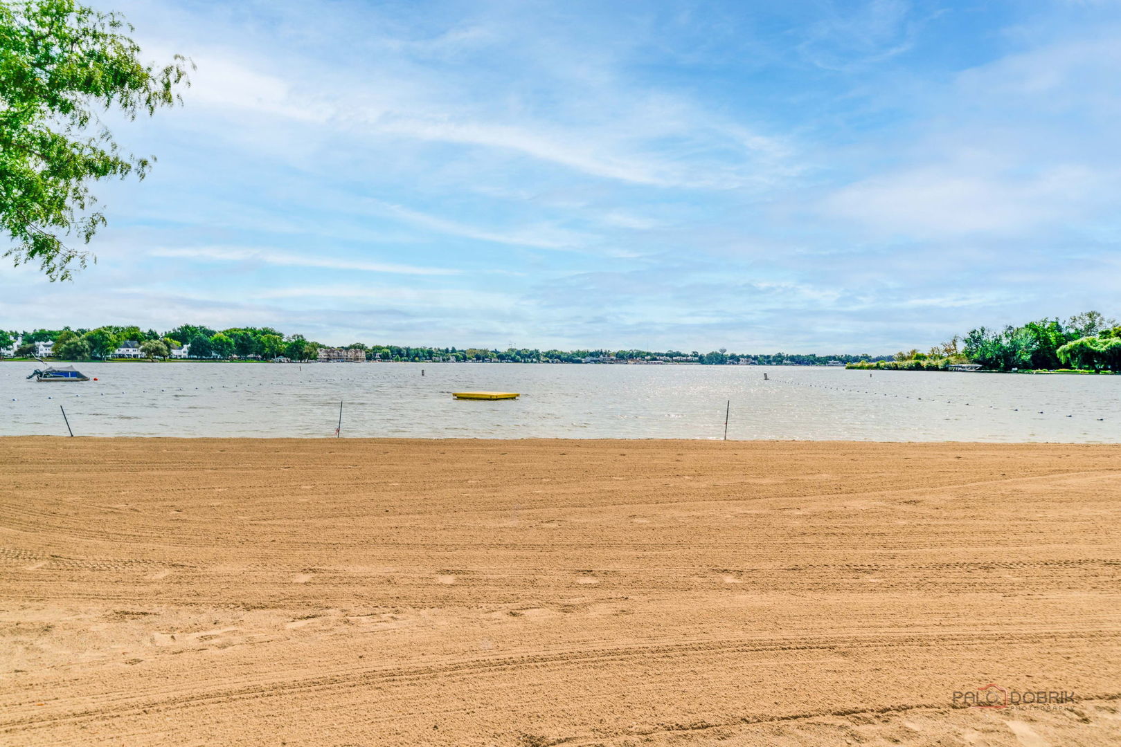Sandy beach shoreline beside a lake, with distant homes and trees across the water.