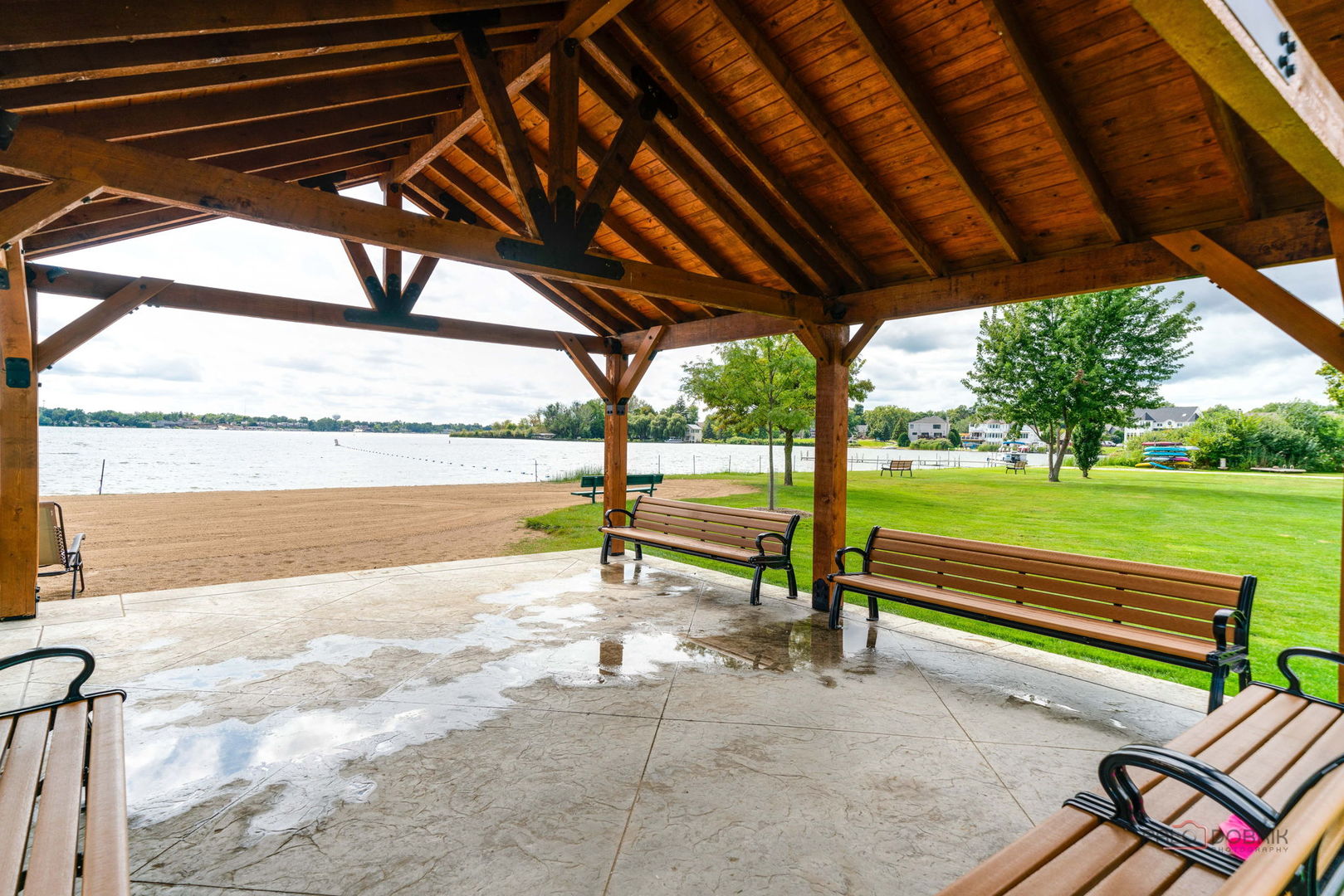 Covered timber pavilion with benches overlooking a lake, sandy beach, and green lawn.