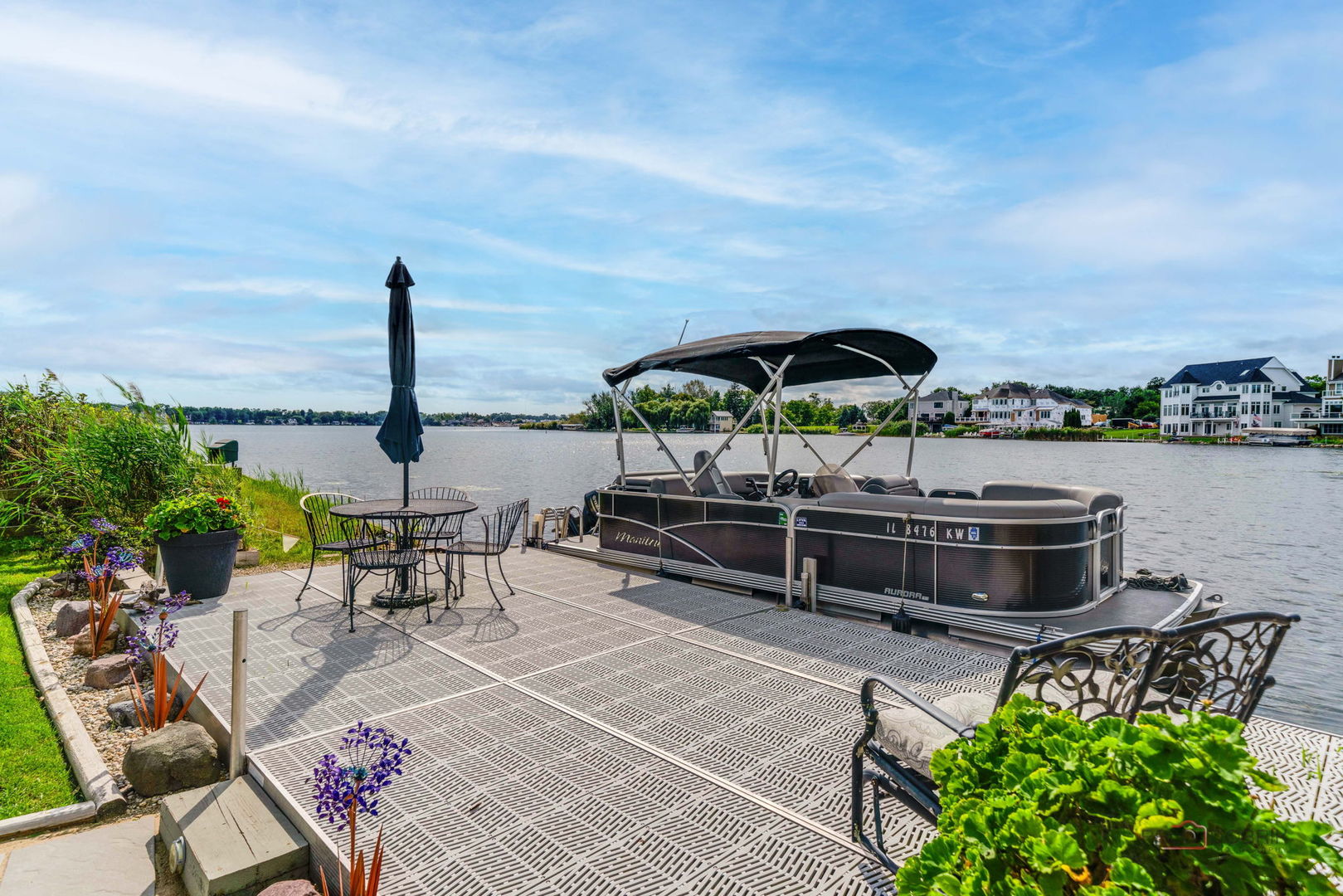 Lakeside dock with a covered pontoon boat, patio seating, and homes across the water.