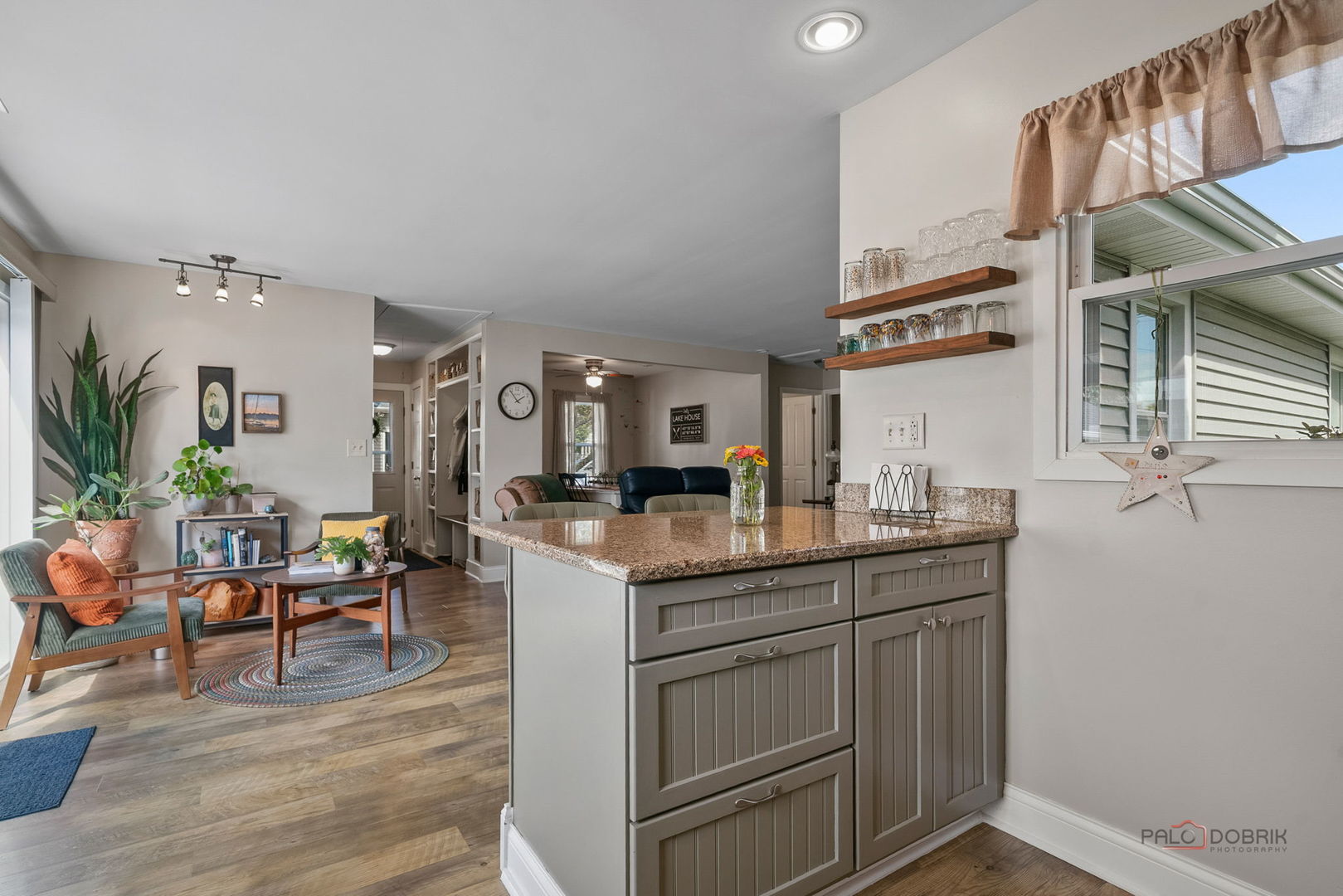 Open-plan kitchen with a granite island and wooden shelves beside a furnished living area.