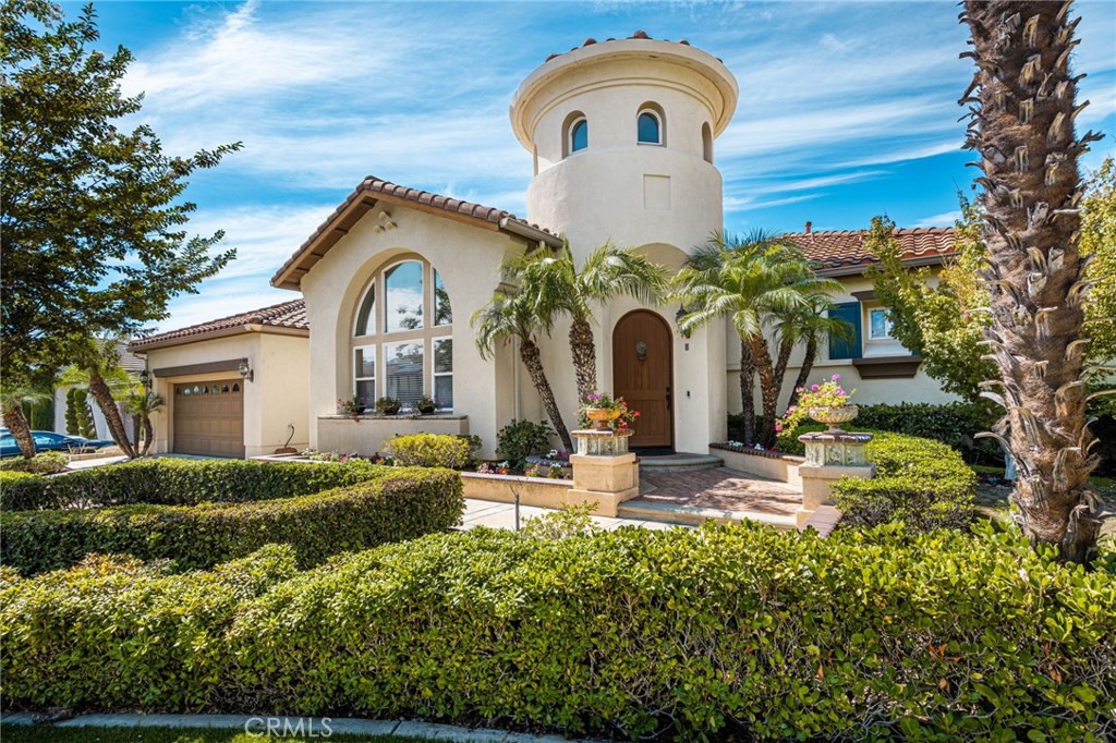 Mediterranean-style home with a round tower, arched window, palm trees, and clipped hedges.