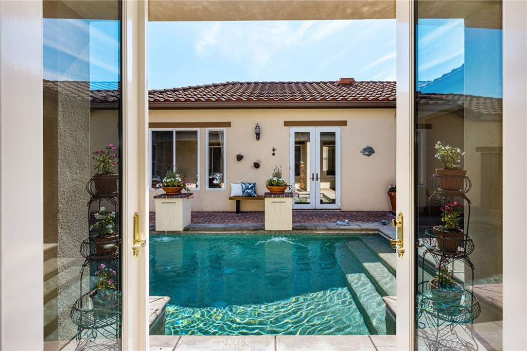 Swimming pool viewed through open glass doors, with a tiled patio and planters beyond.
