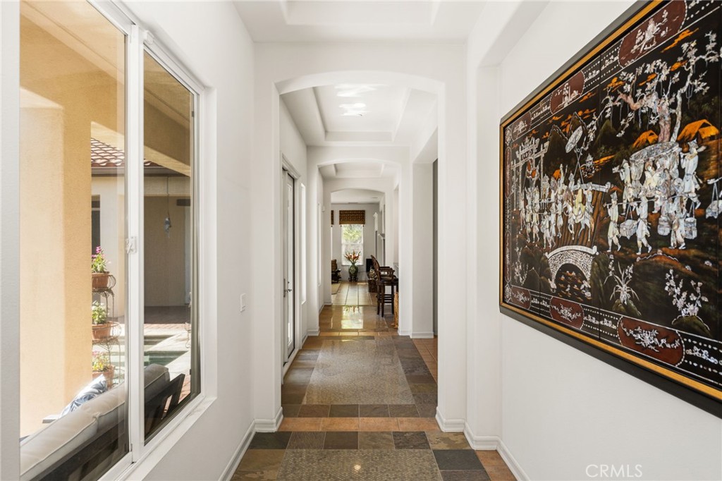 Long tiled hallway with white walls, a large framed artwork, and windows overlooking a courtyard.