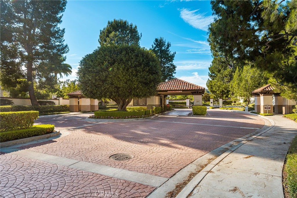Gated community entrance with tiled-roof guard booths, mature trees, and a brick-paved drive.