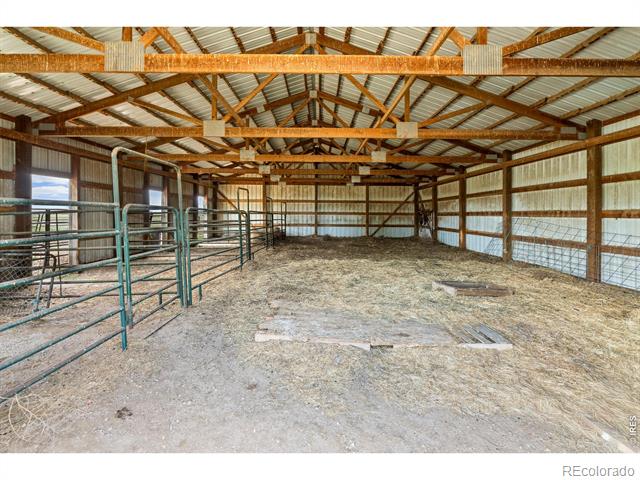 Barn interior with metal livestock gates, a dirt-and-straw floor, and exposed wooden roof trusses.