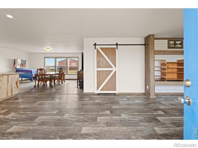 Open-plan interior with gray plank flooring, a white barn door, and a dining area by large windows.