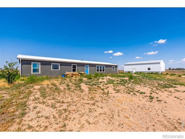 Single-story dark-sided home with a blue front door and detached metal outbuilding on a sandy lot.