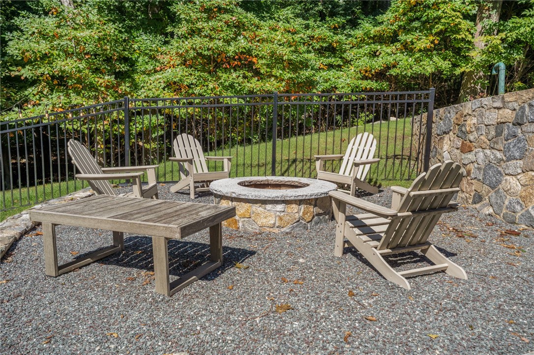 Gravel patio with Adirondack chairs, a stone fire pit, and a black metal fence amid trees.