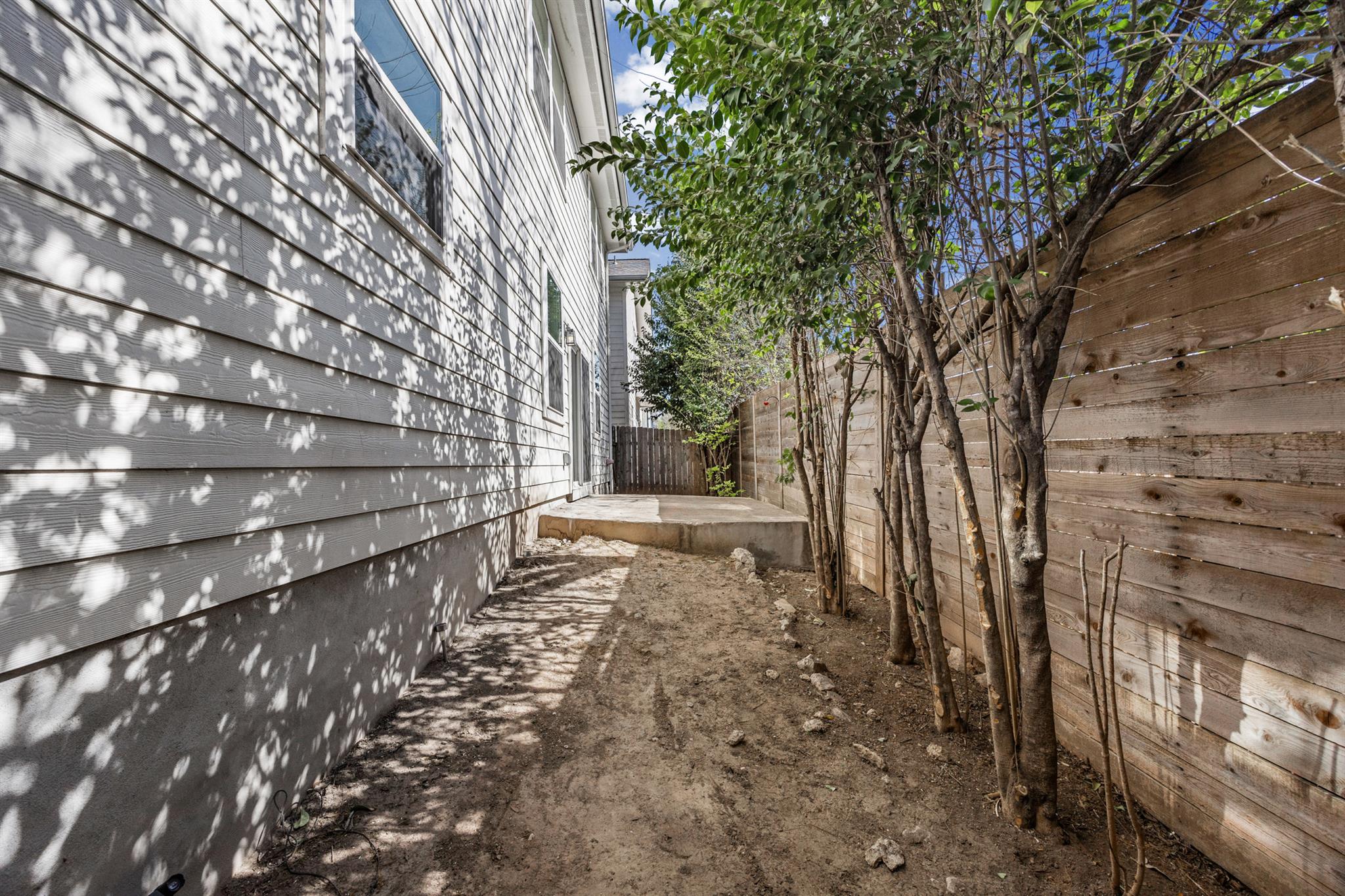 Narrow dirt side yard with young trees, a wooden privacy fence, and a small concrete patio.