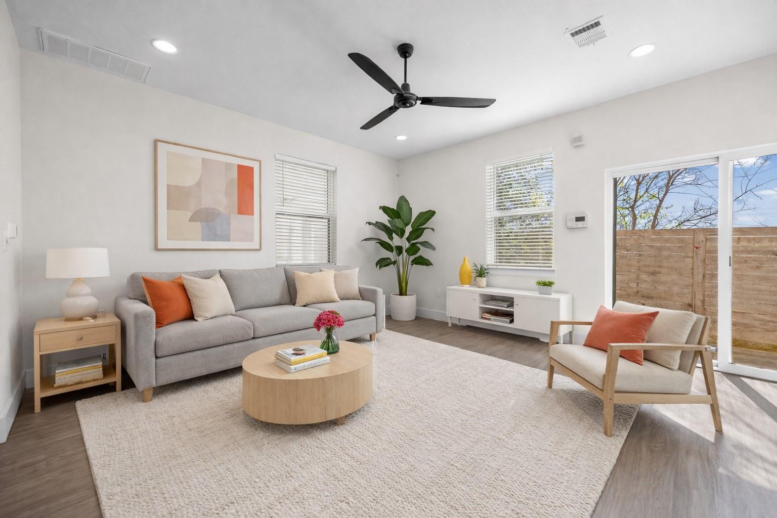 Staged living room with gray seating, a light wood coffee table, ceiling fan, and sliding glass door to a fenced patio.