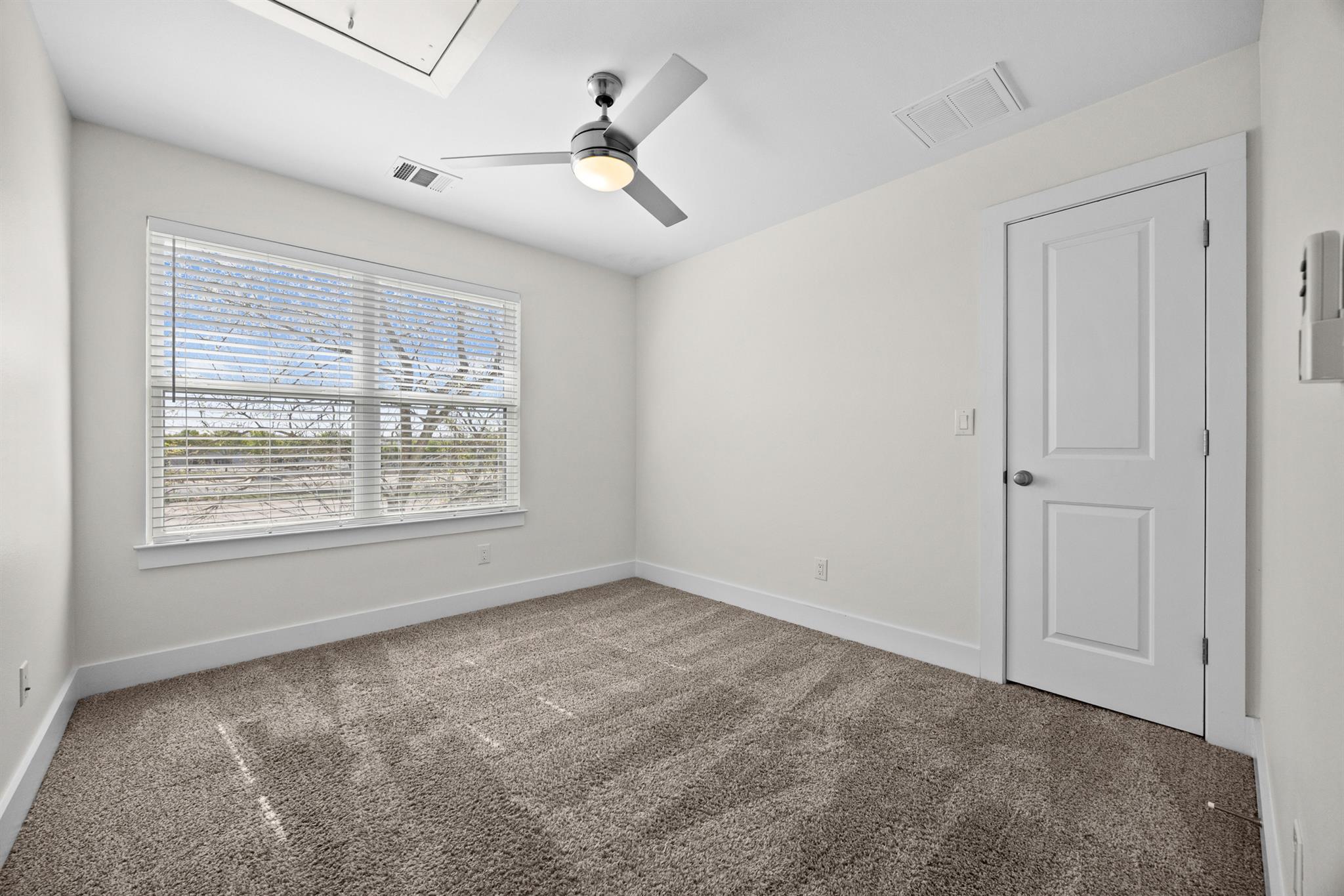 Empty bedroom with beige carpet, a wide window with blinds, and a ceiling fan.