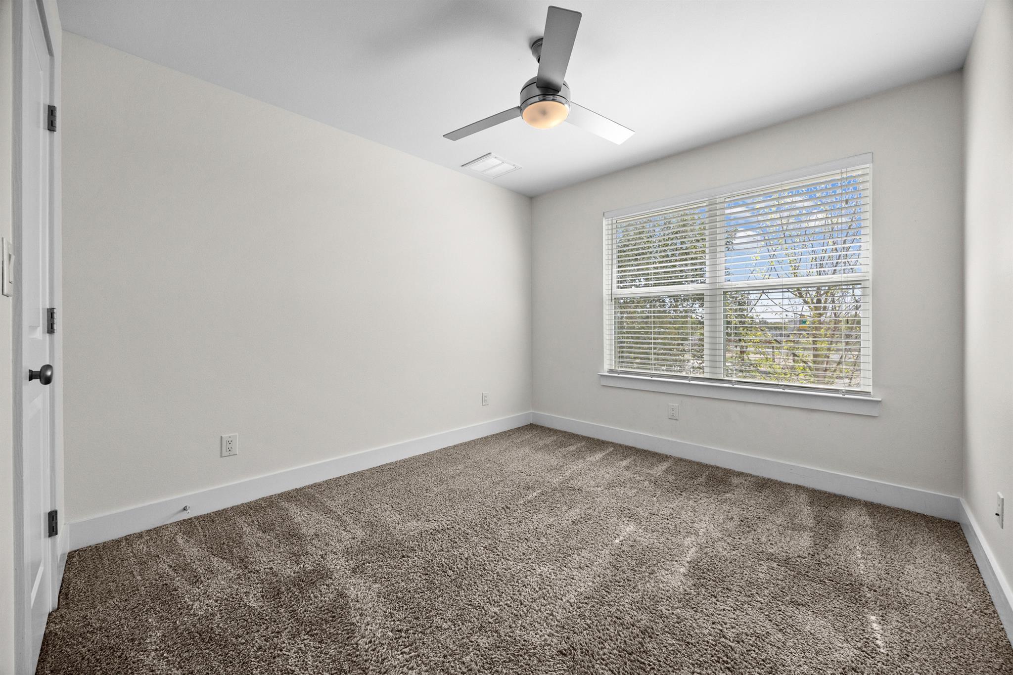 Empty bedroom with beige carpet, a ceiling fan, and a large window with blinds.