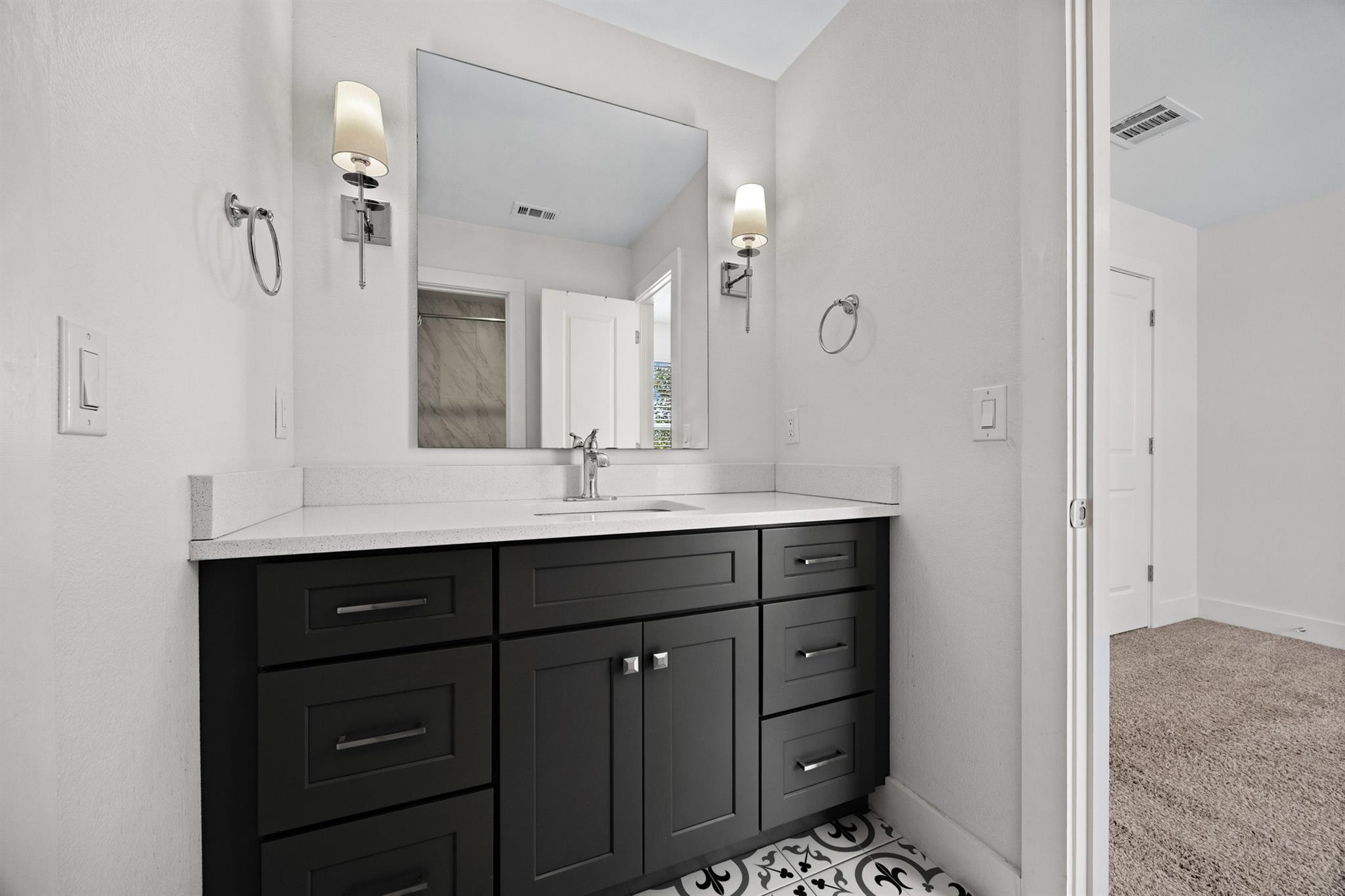 Bathroom vanity with dark cabinets, a wide mirror, wall sconces, and patterned tile floor.