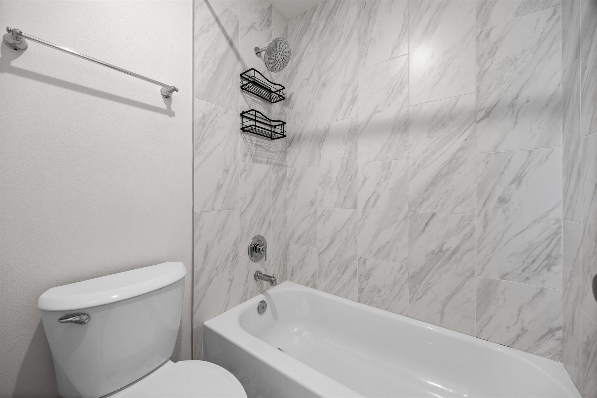 Bathroom with a white tub, gray-veined tile surround, chrome showerhead, and toilet.