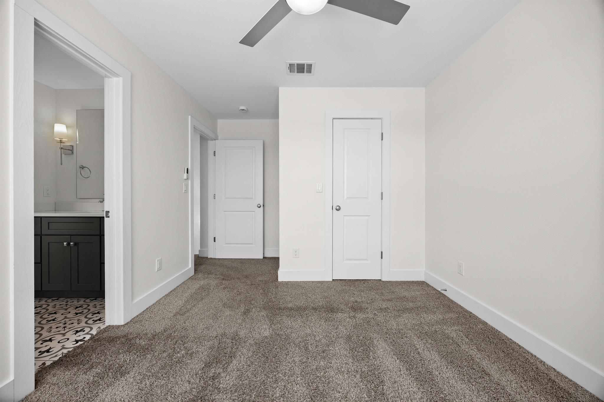 Empty carpeted bedroom with ceiling fan and open doorway to a bathroom with patterned tile.