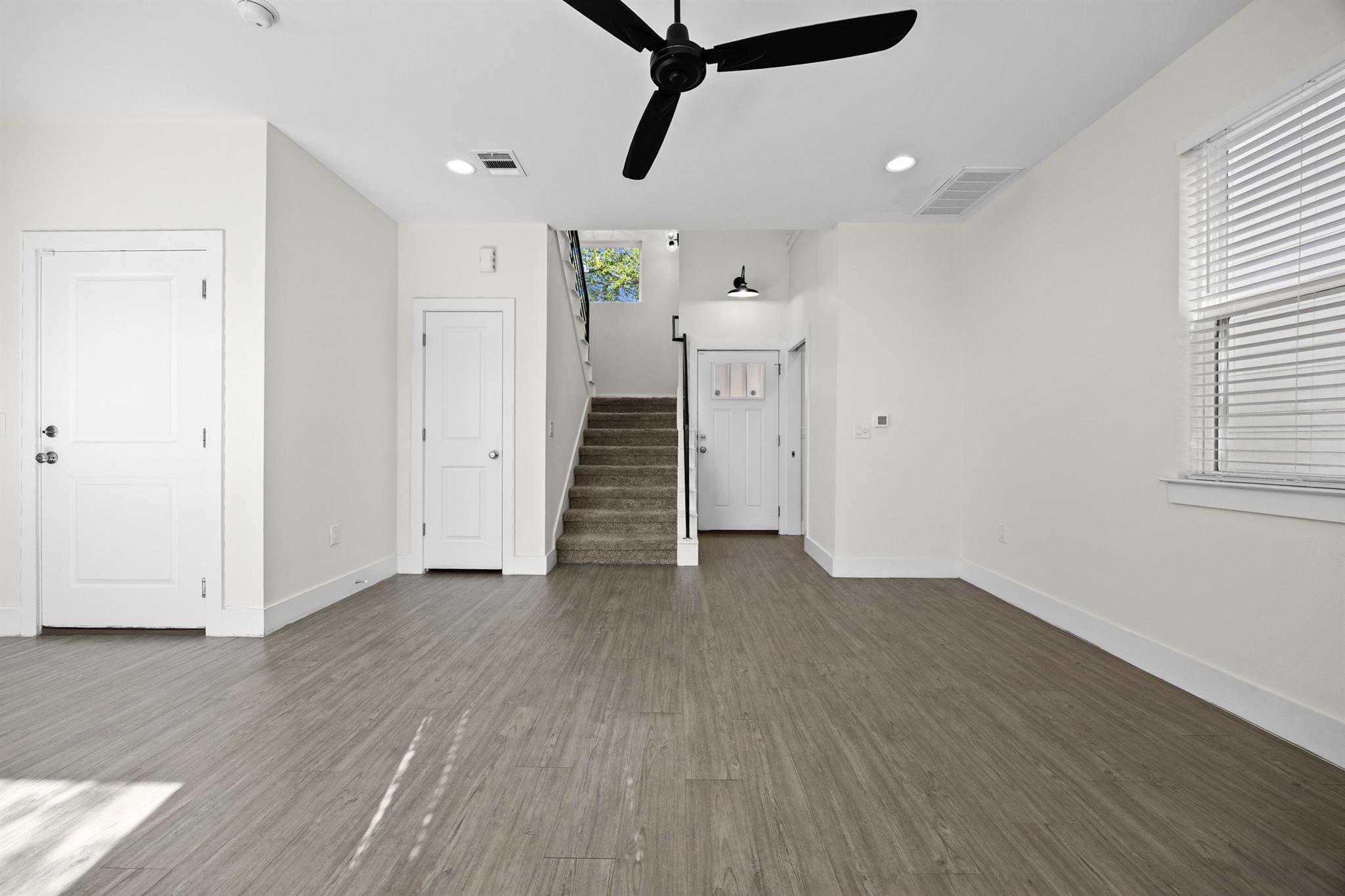 Vacant interior with gray wood-look floors, a carpeted staircase, ceiling fan, and front door.