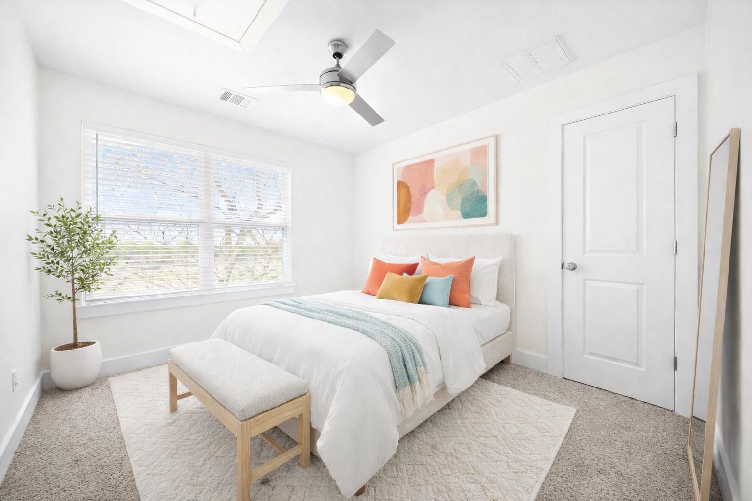 Staged bedroom with a white bed, colorful accent pillows, and a large window with blinds.