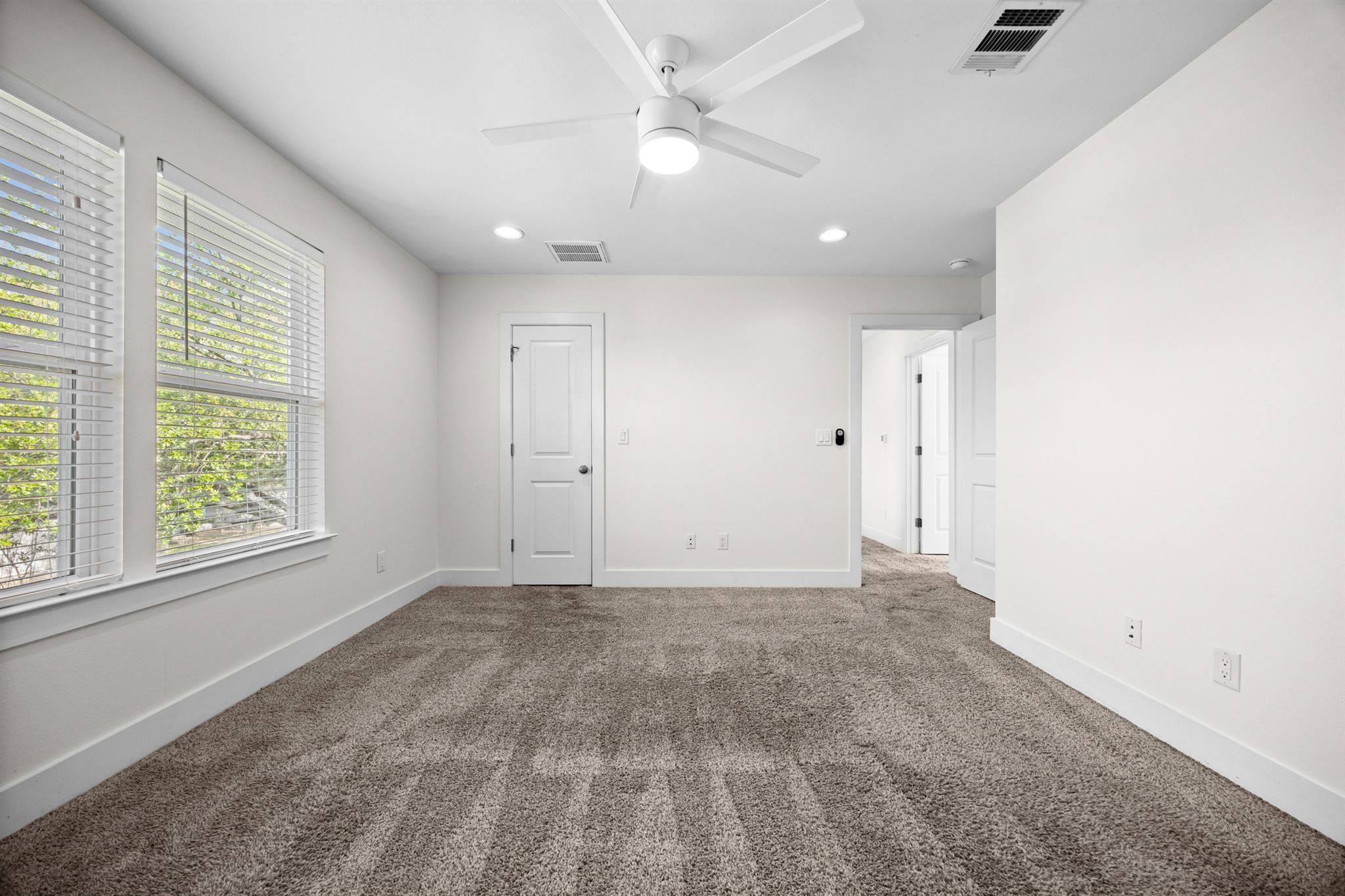 Empty bedroom with gray carpet, two windows with blinds, and a ceiling fan.