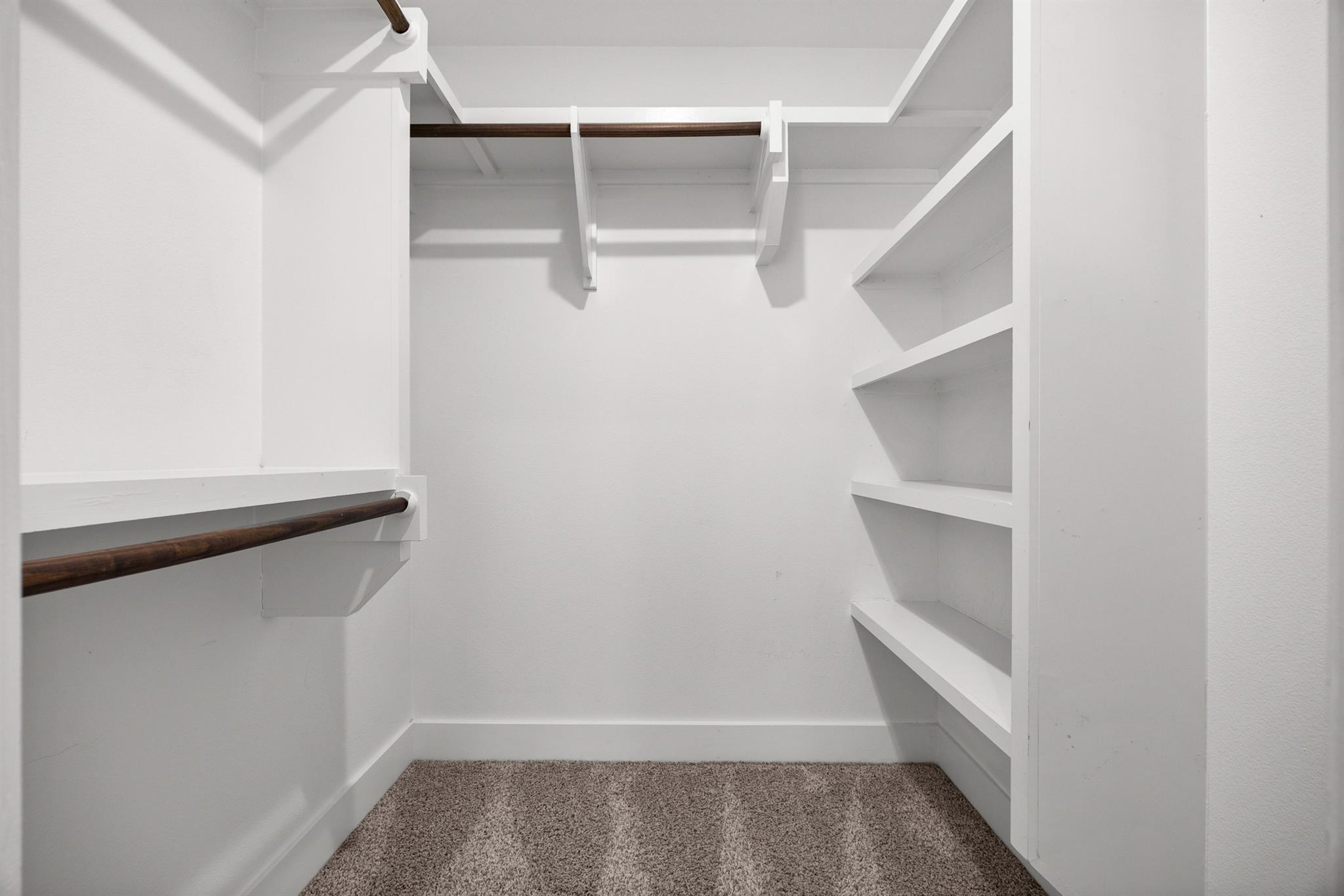 Walk-in closet with built-in white shelves, wooden hanging rods, and beige carpet.