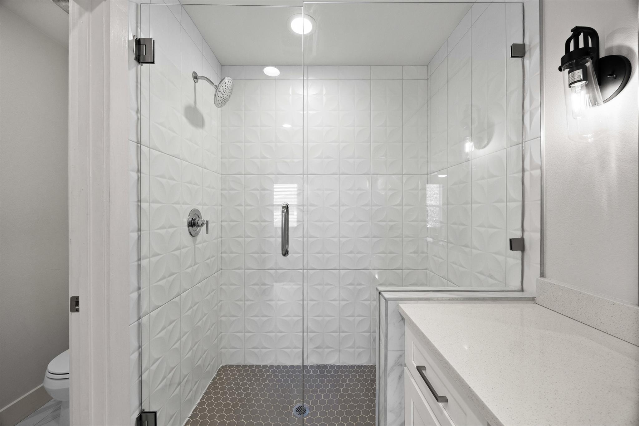 Bathroom with a glass-enclosed shower, sculpted white tile, dark hexagonal floor, and white vanity.