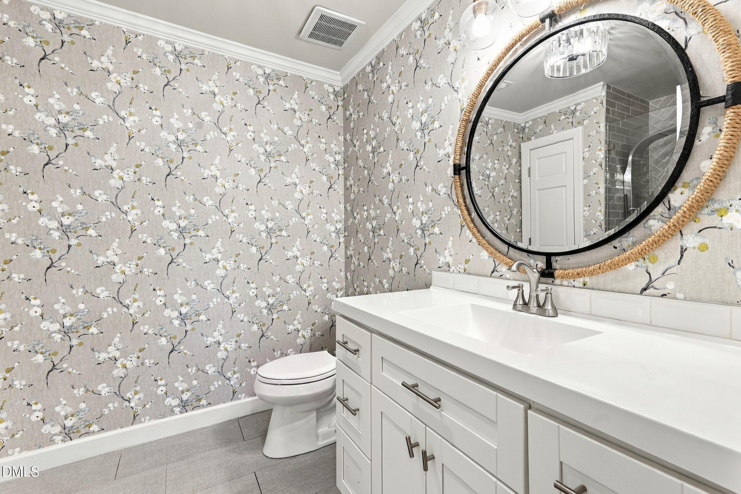 Bathroom with white vanity, floral wallpaper, and a large round mirror with a woven frame.