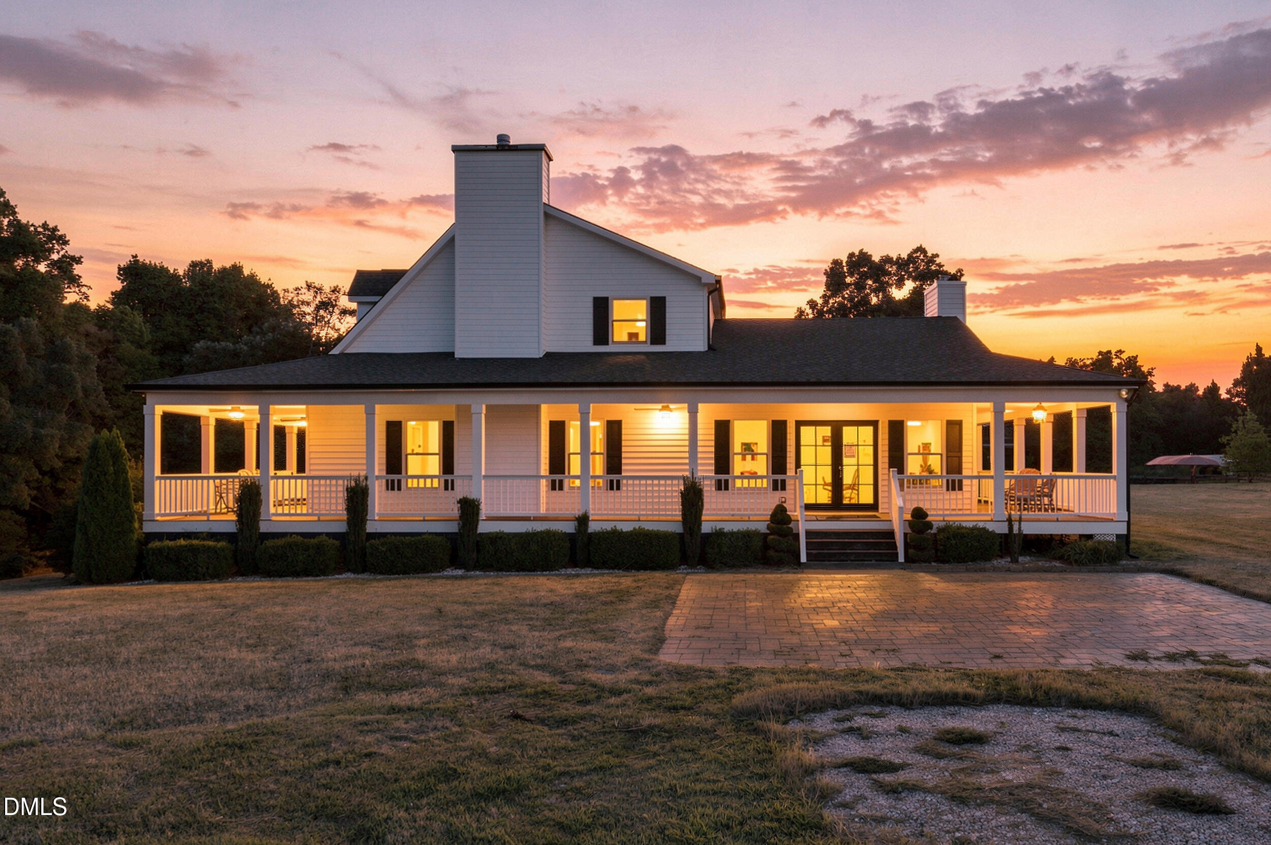 White two-story house with an illuminated wraparound porch and brick driveway at sunset.
