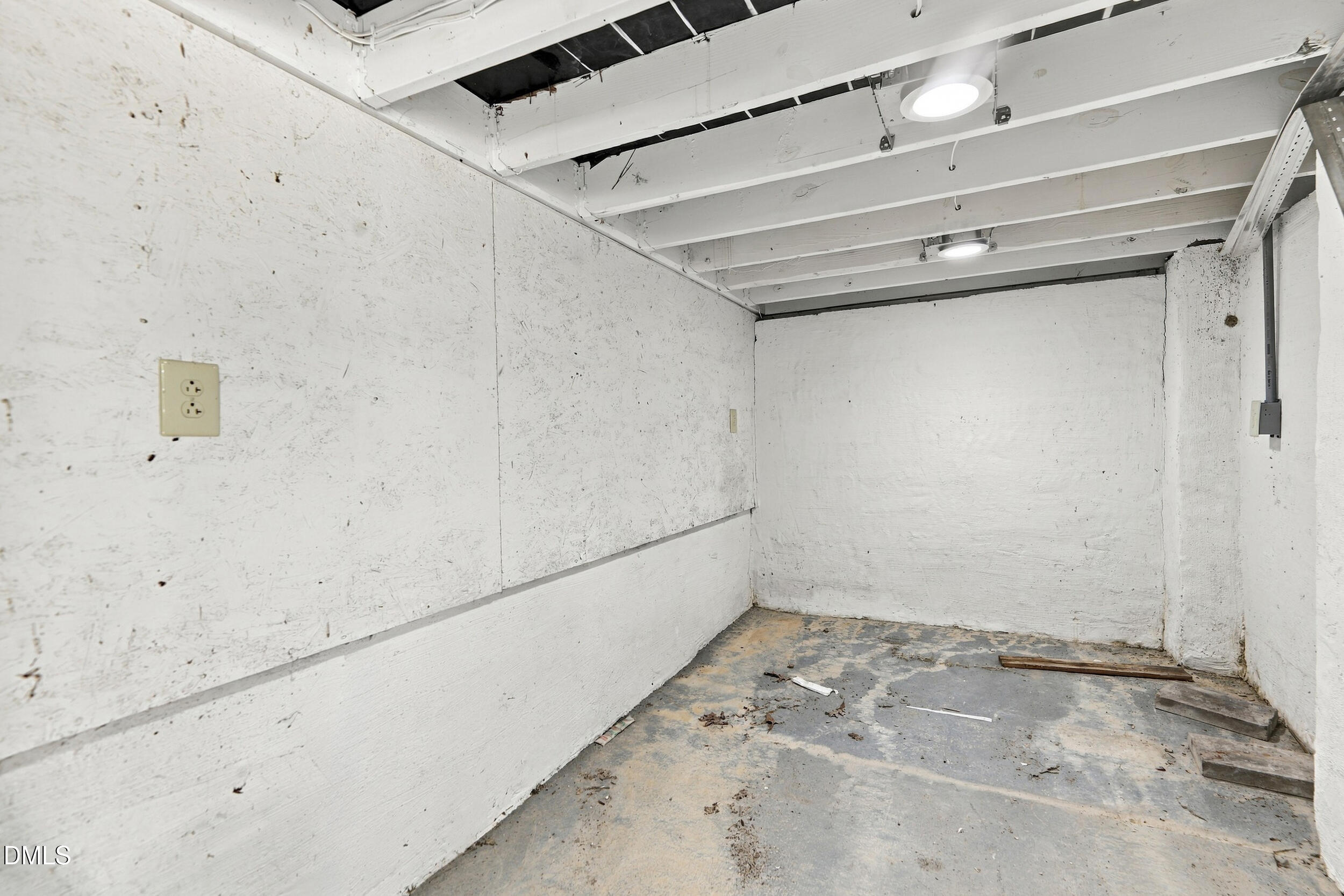 Empty unfinished garage with scuffed white walls, exposed ceiling beams, and debris on the concrete floor.