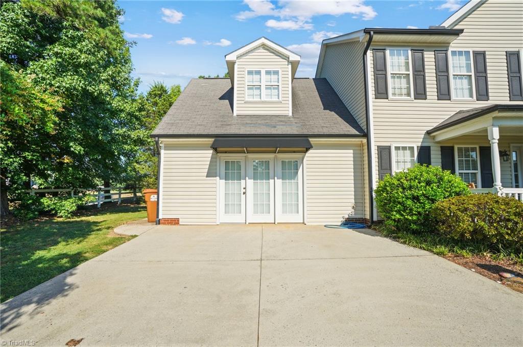 Two-story light-colored house with dark shutters, a broad driveway, and mature trees.