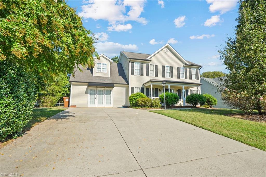 Two-story beige home with a broad concrete driveway, covered porch, and mature trees.