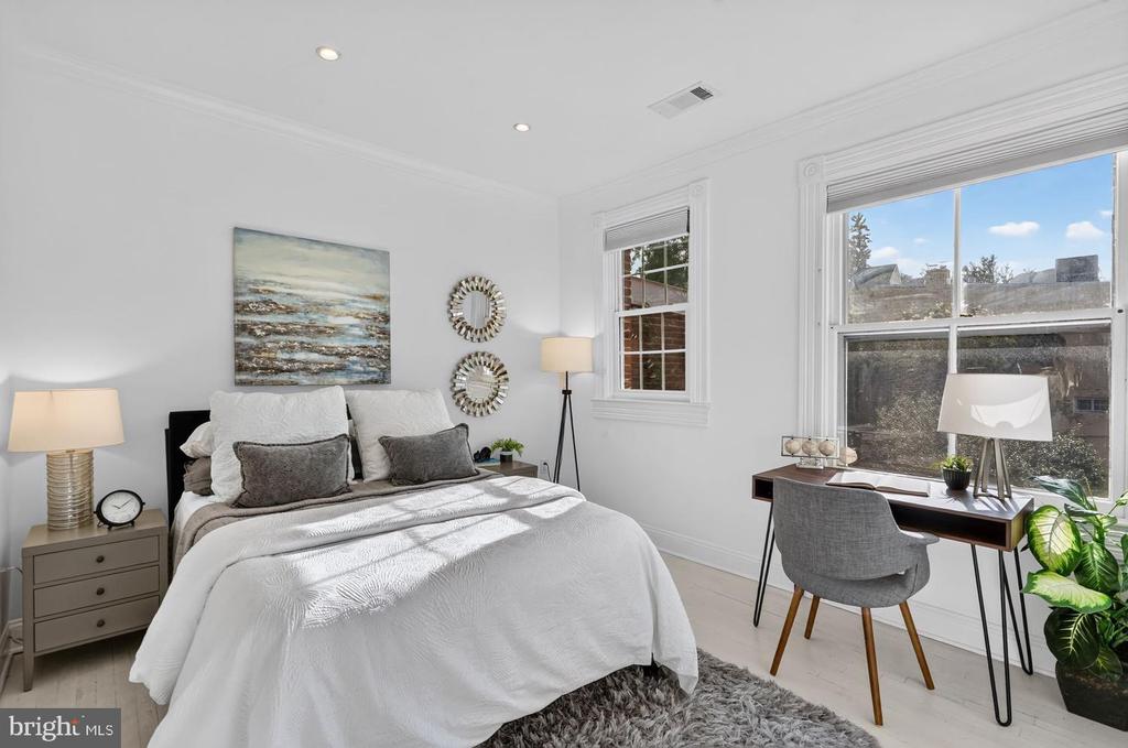 Staged bedroom with a white bed and a desk beside two large windows.