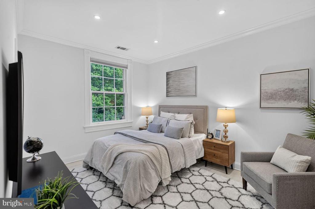 Staged bedroom with a gray upholstered bed, large window overlooking greenery, and a gray armchair.