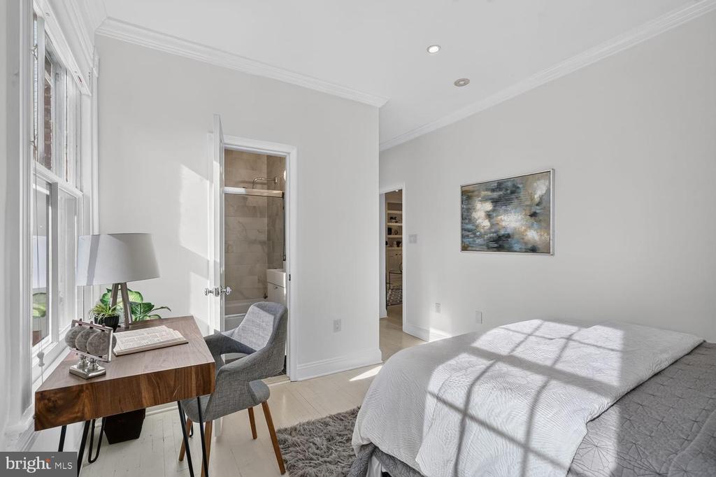Bright bedroom with a white bed, large windows, wooden desk, and doorway to a tiled bathroom.