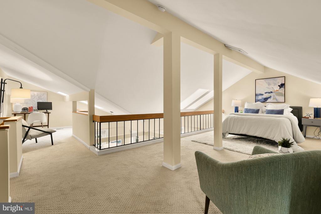 Loft bedroom with a king bed, sloped ceiling, and carpeted sitting area beside the railing.