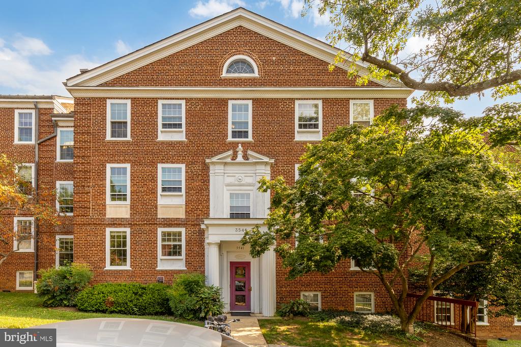 Red-brick apartment building with white-trimmed entrance, mature trees, and landscaped grounds.