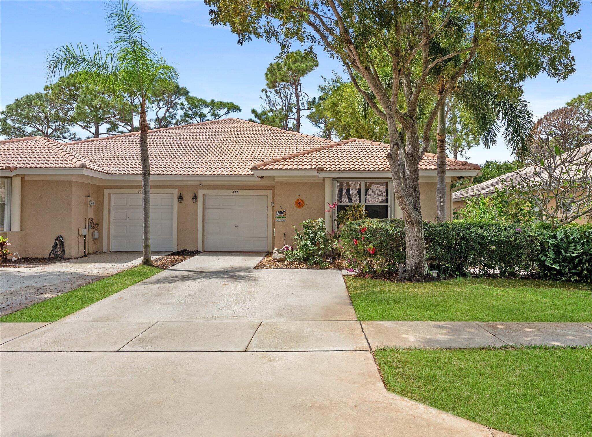 Beige stucco home with a tile roof, two garage doors, and a wide driveway beneath mature trees.