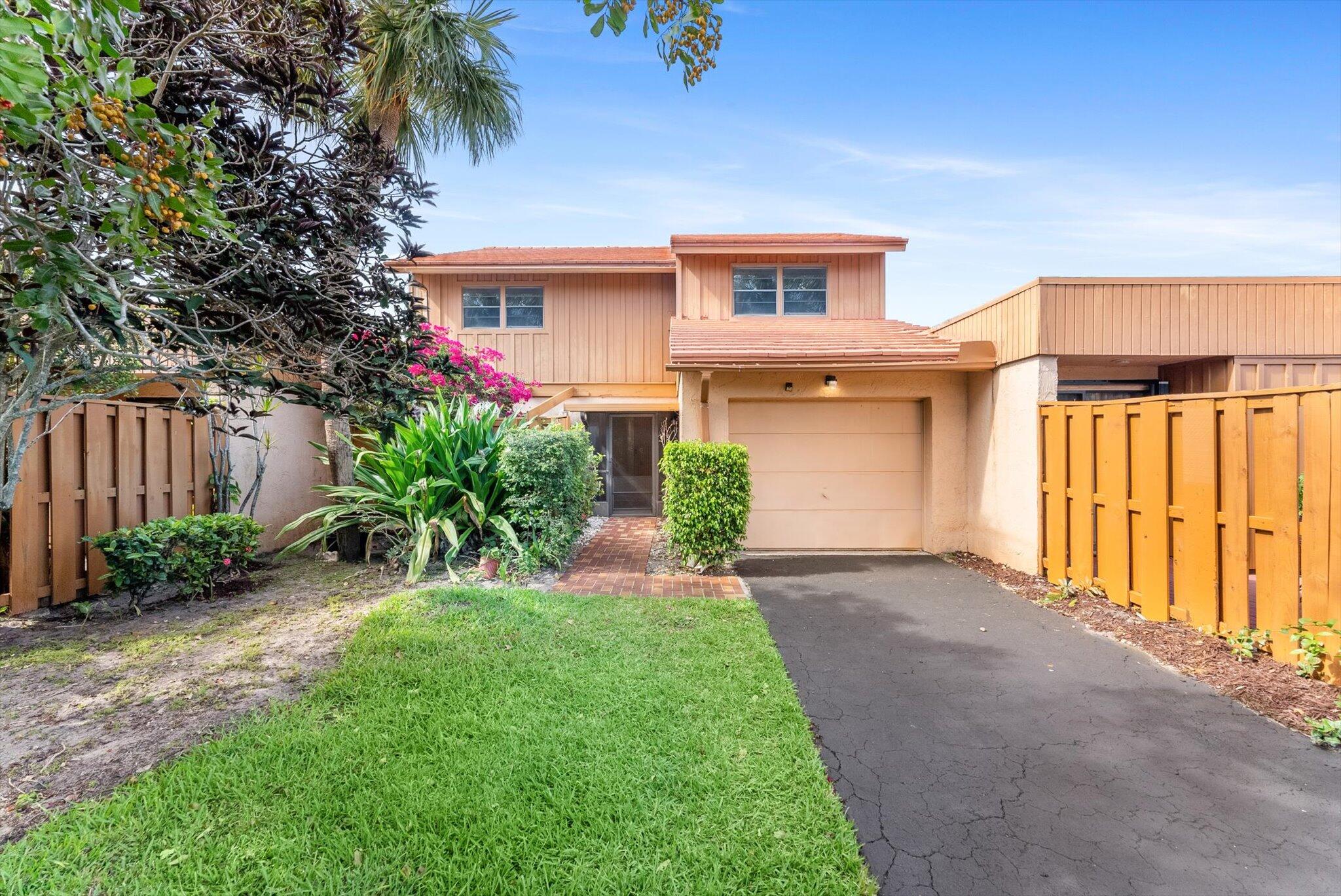 Tan two-story home with a garage, tropical landscaping, and a broad driveway.