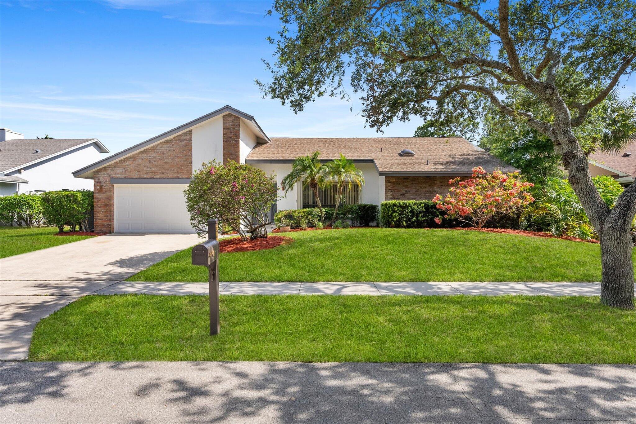 Single-story brick and stucco home with an attached garage, flowering shrubs, and a large shade tree.