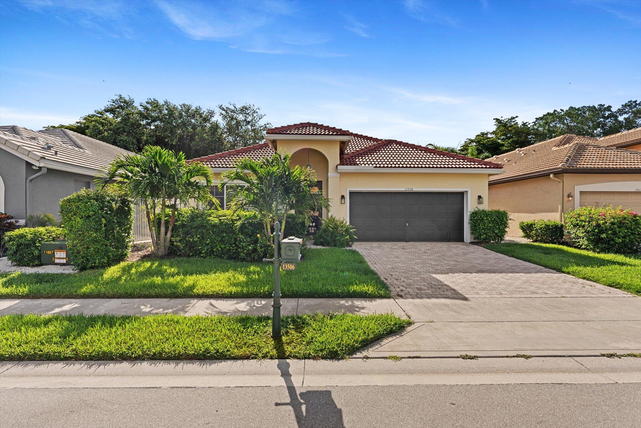 Tan stucco home with a red tile roof, dark double garage door, tropical landscaping, and paved driveway.
