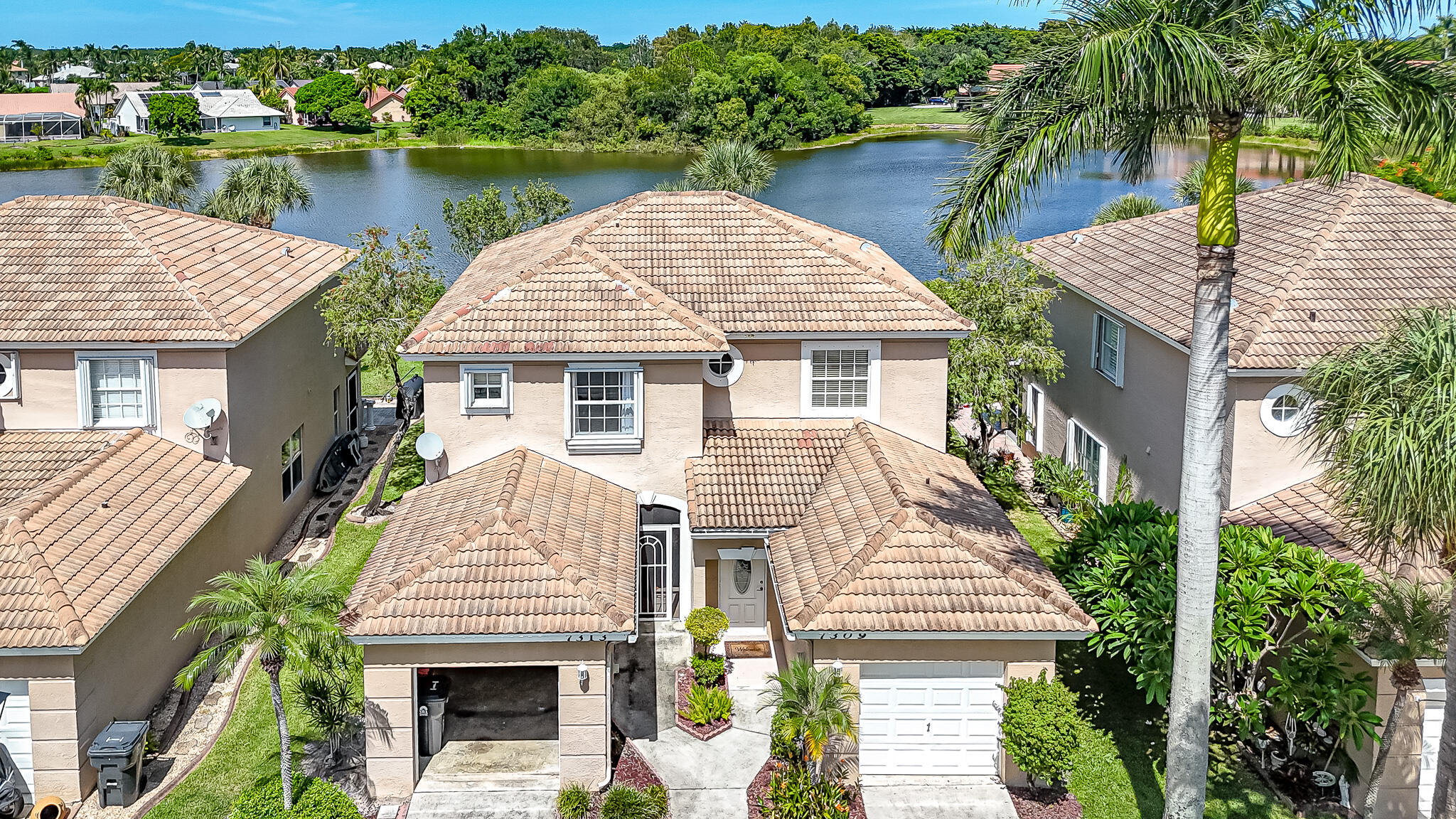 Aerial view of a tile-roofed home beside a lake, surrounded by neighboring houses and palm trees.