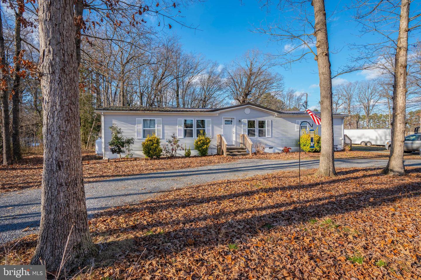 Single-story home with light siding, front steps, gravel driveway, and a wooded, leaf-covered yard.