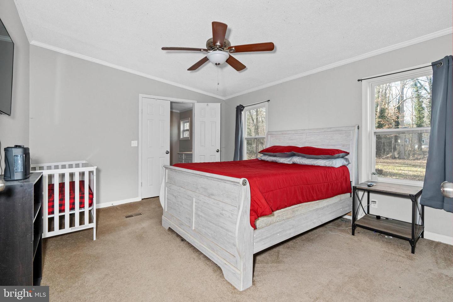 Bedroom with a white bed in red bedding, a white crib, and two curtained windows.