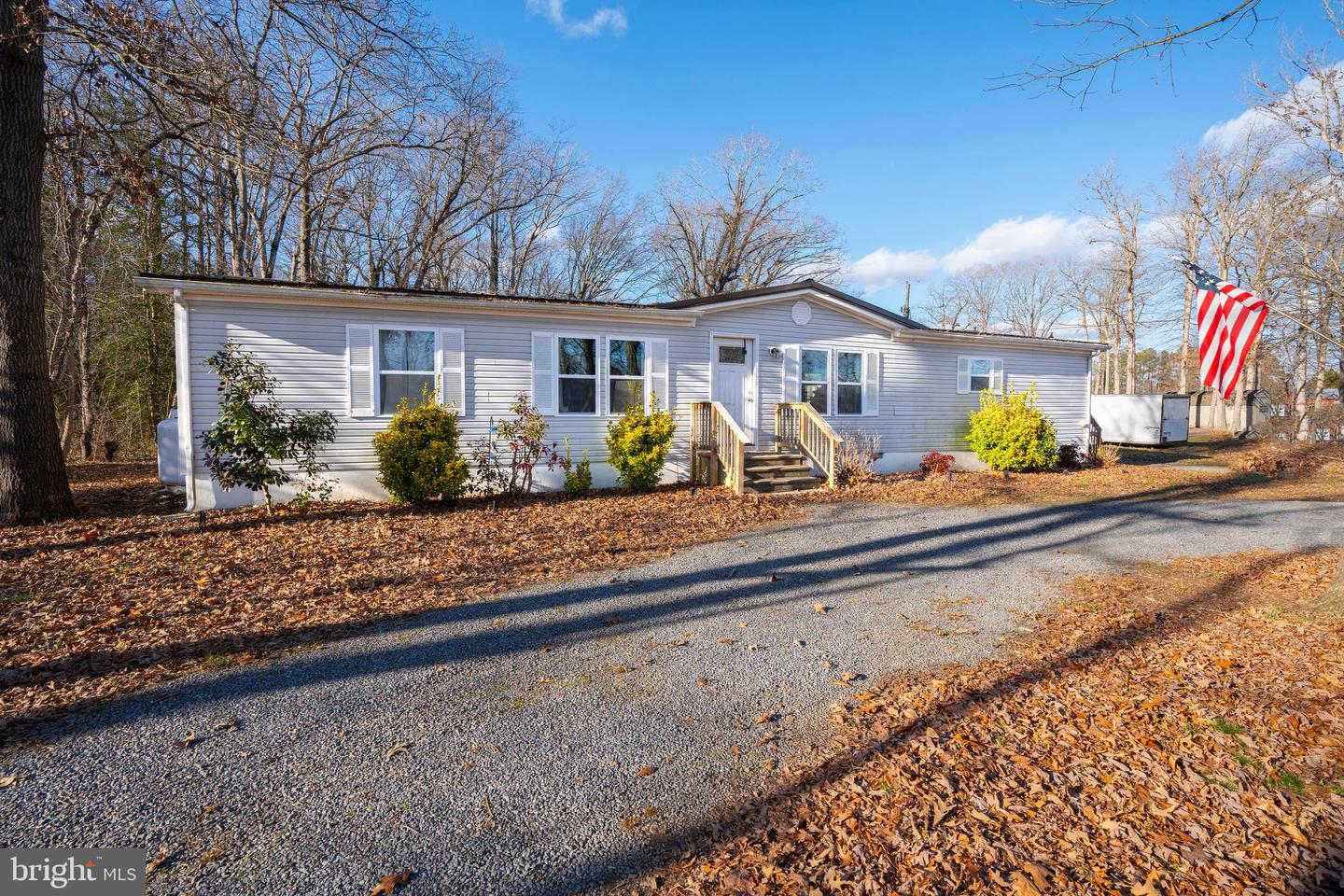 Single-story gray home with white trim, front steps, and an American flag beside a gravel drive.