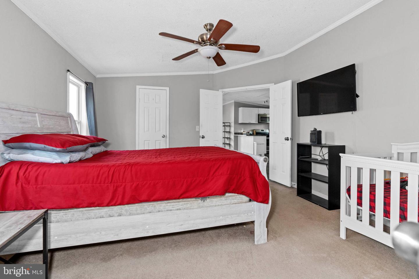 Bedroom with a red-quilted bed, white crib, ceiling fan, and open doors to the kitchen.